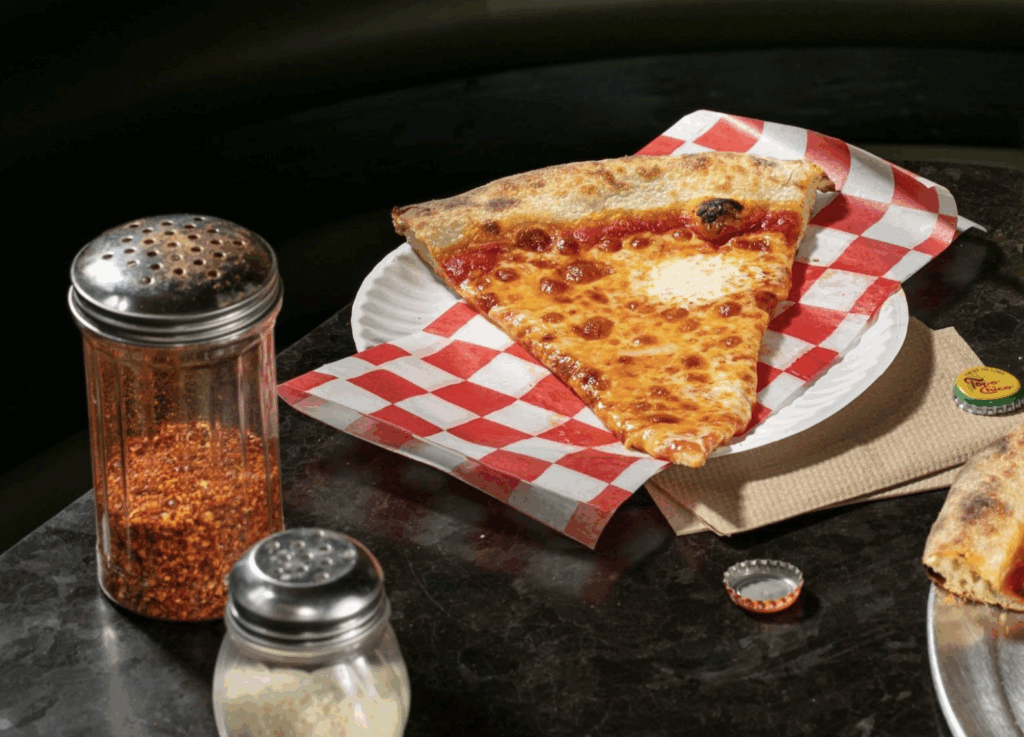 Upside Pizza slice on a metal tray with parmesan and red pepper shakers — one of the most popular pizza places in Manhattan for late-night slices.